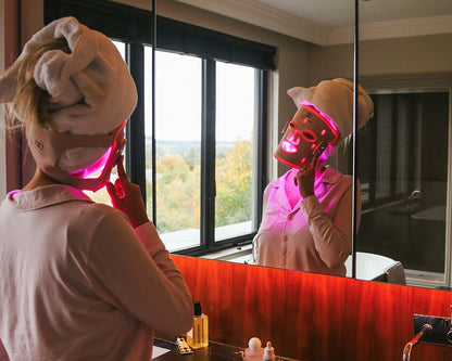 Person wearing a pink LED face mask in front of a mirror with a scenic view.