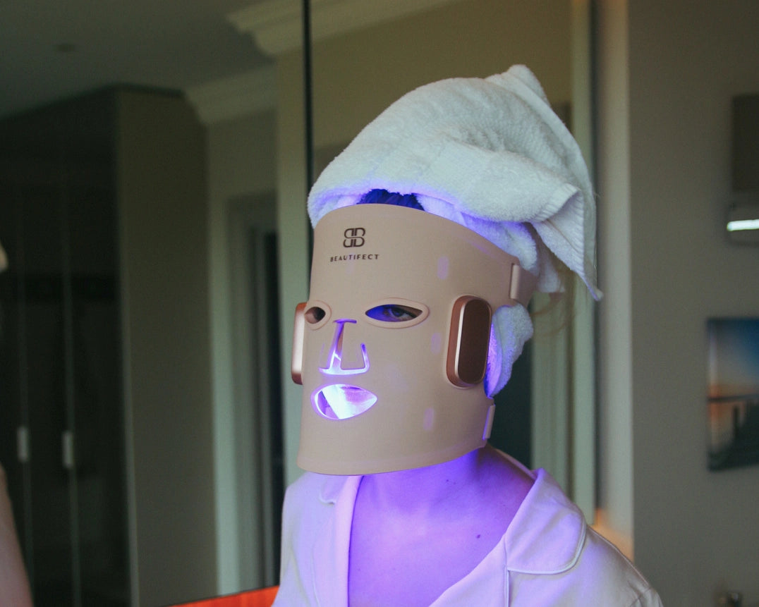 Person wearing a LED face mask with a towel on their head in a room setting.