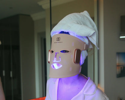 Person wearing a LED face mask with a towel on their head in a room setting.