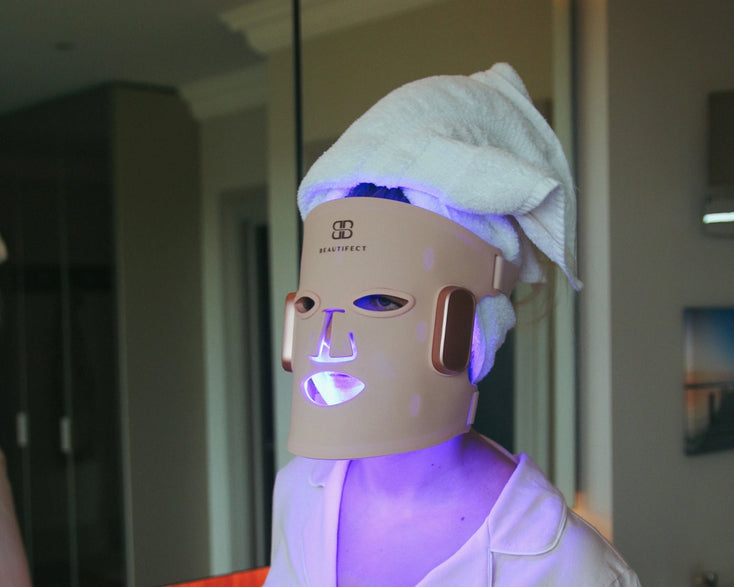 Person wearing a LED face mask with a towel on their head in a room setting.
